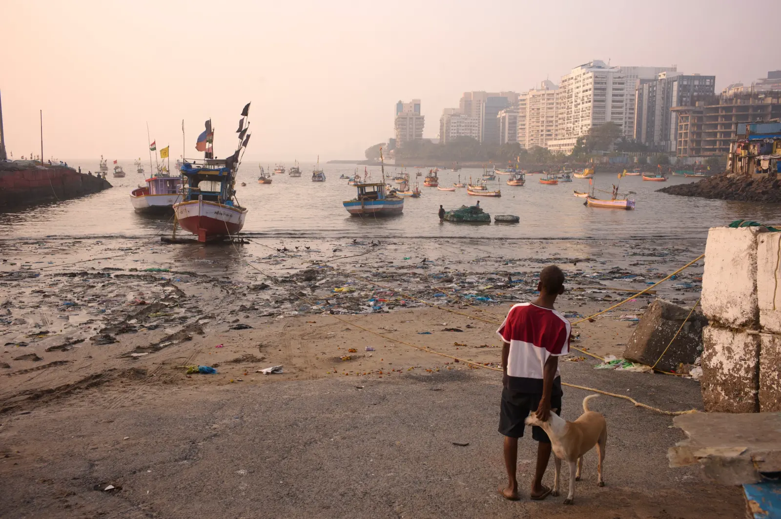 Fishing boats in a polluted harbour with high-rise buildings on the far shore and a man — From Mumbai to Ahmedabad, Urban Transects — Mumbai, Ahmedabad, India.