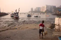 Fishing boats in a polluted harbour with high-rise buildings on the far shore and a man — From Mumbai to Ahmedabad, Urban Transects — Mumbai, Ahmedabad, India.